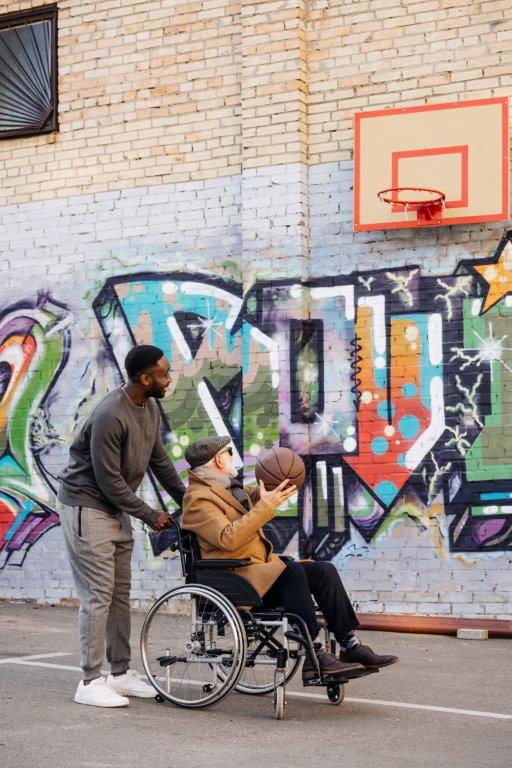 En yngre man som kör en äldre man i rullstol med basketboll framför en grafittivägg med basketkorg