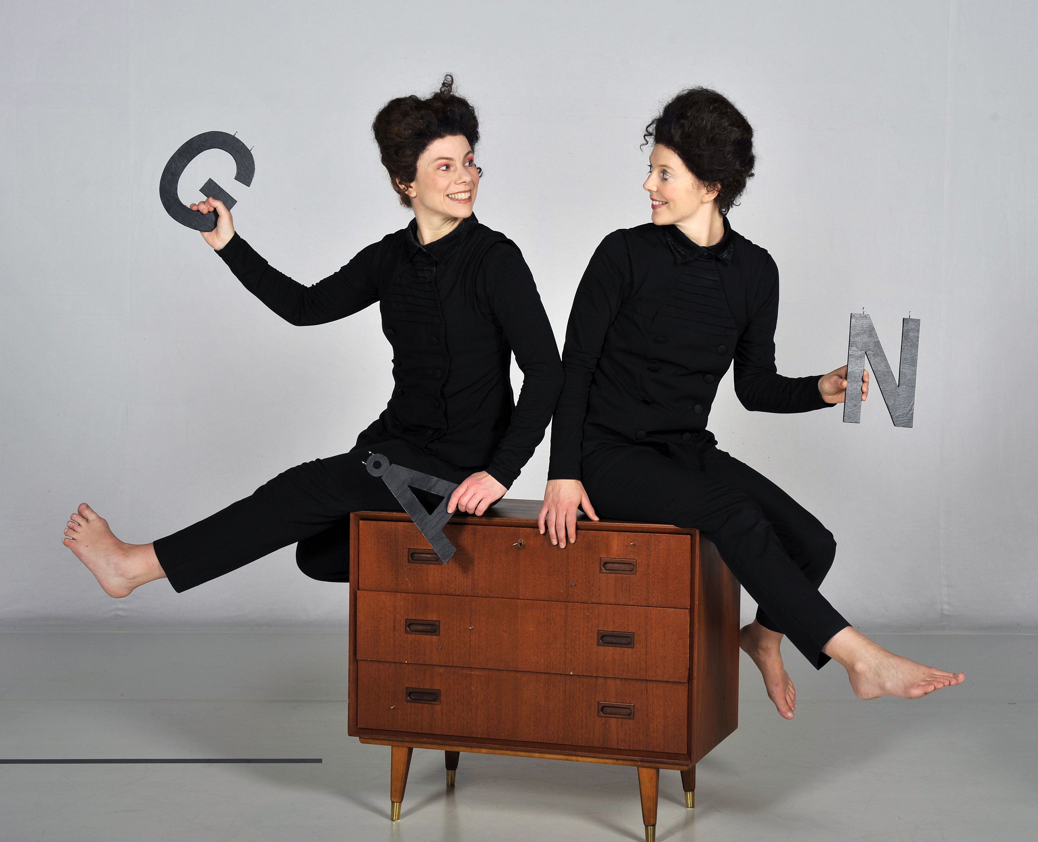 Två svarklädda dansare med bokstäver i händerna sitter på en byrålåda