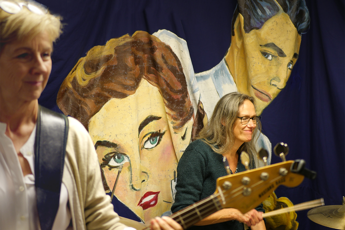 Eva Grund med långt hår och glasögon spelar trummor. Lena Andersson med kort ljust hår spelar bas. Båda mot en blå bakgrund.