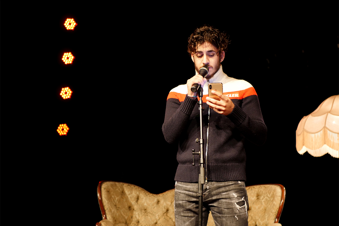 Poeten Ali Junior står i strålkastarljus på scenen och läser en dikt från mobiltelefonen.