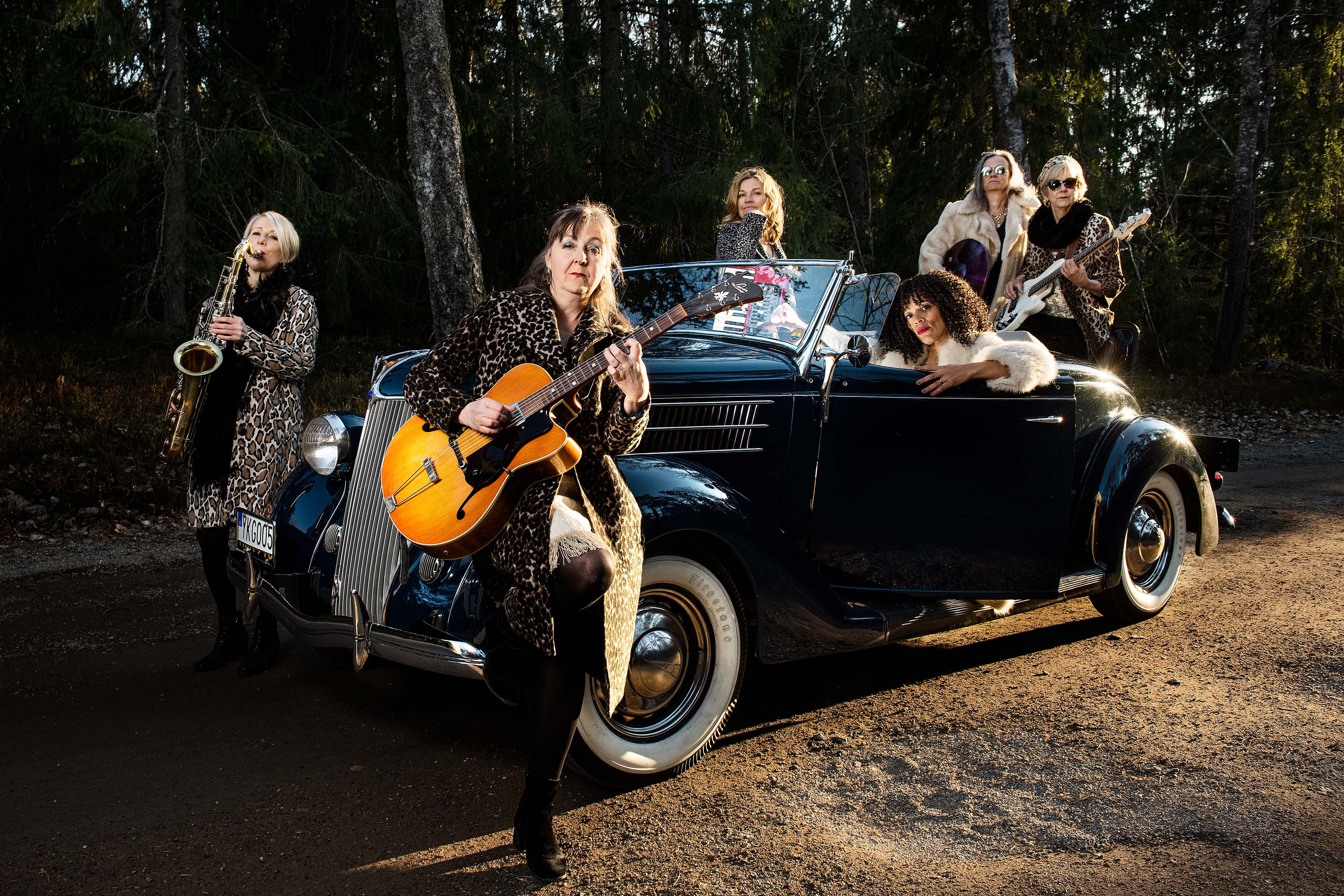 En bild på flera kvinnor i och runt en gammal bil, de håller i instrument och tittar in i kameran